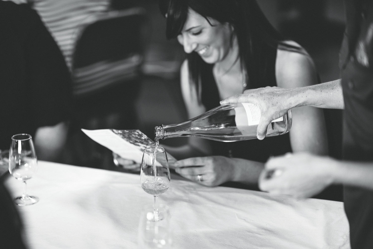 A monochrome image of a woman smiling broadley in anticipation of the glass of cider which is being poured for her at a tasting in Small Acres Cyder cellar door