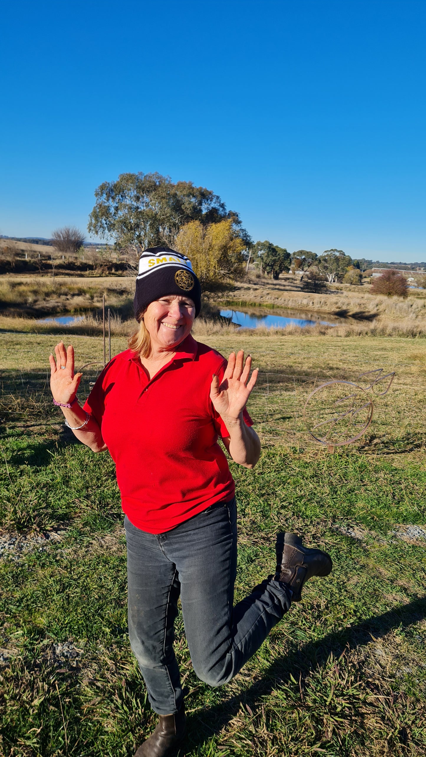 A woman in a redshirt skips while wearing a black, white and gold Small Acres Cyder beanie. The background includes a dam and some trees.