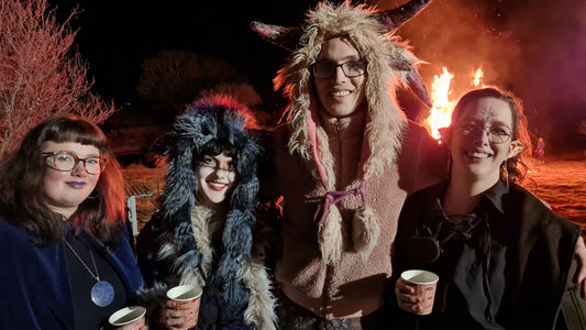 A group of revellers in medieval costume hold cups of warm cider with a roaring fire in the background.