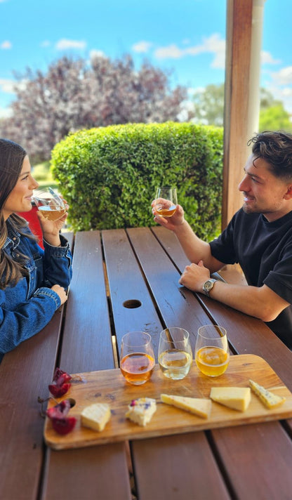 A young couple at a picnic table with a selection of cheeses and ciders. The woman is about to taste.