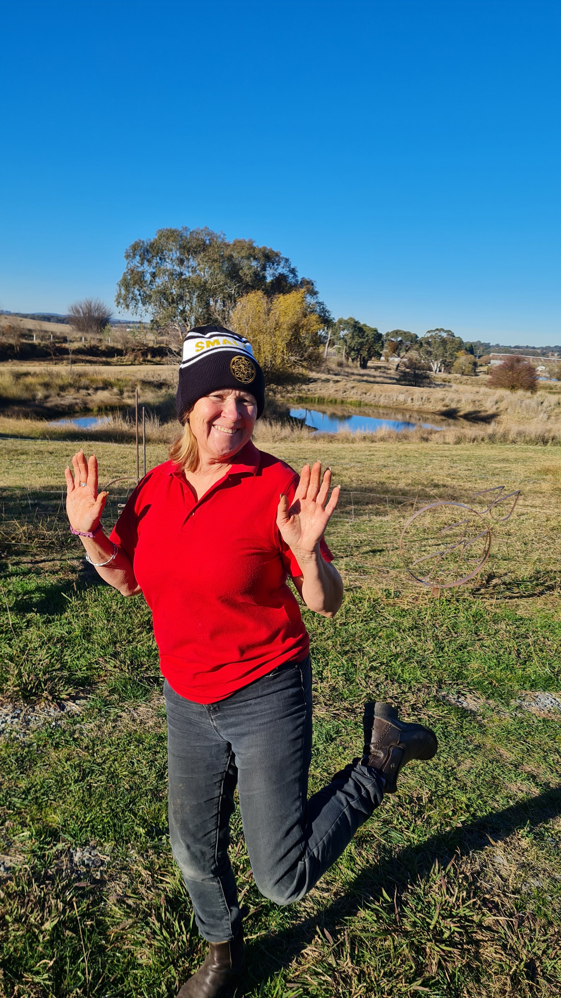 A woman in a redshirt skips while wearing a black, white and gold Small Acres Cyder beanie. The background includes a dam and some trees.