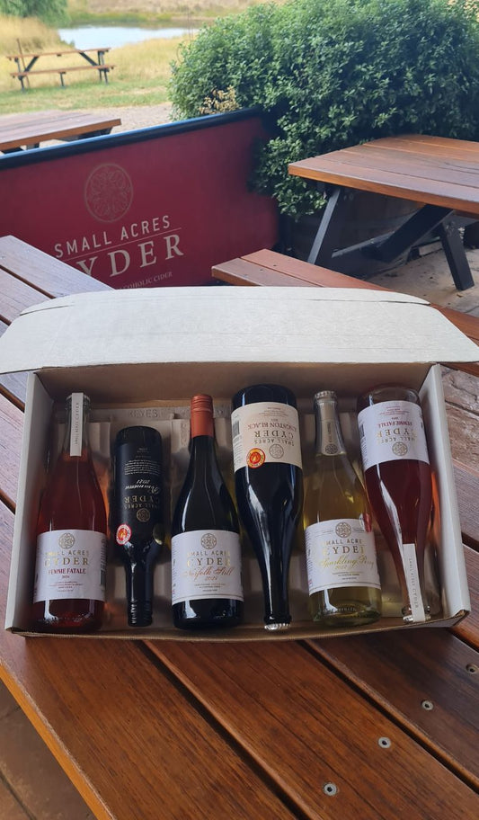 Box of cider on a wooden picnic table including 2 x Femme Fatale, a Pommeau, a Norfolk Still, a Kingston Black, a Sparkling Perry