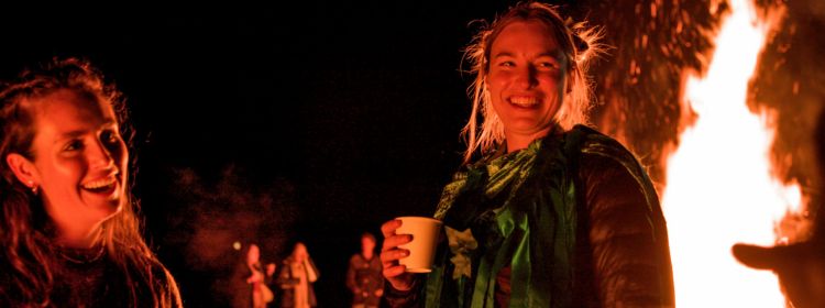 Two women enjoying cider in front of a large burning fire at the Wassail Event