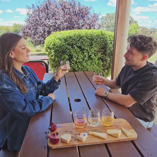 A couple clink glasses sitting at a picnic table with blue skies and a wild plum tree in the background