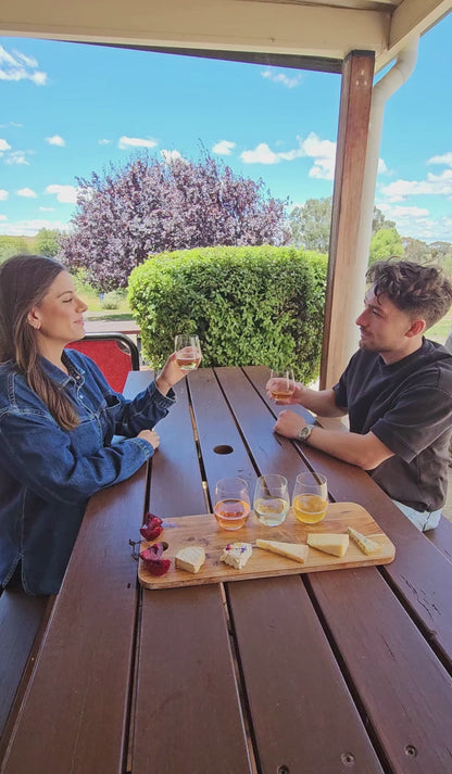 A couple clink glasses sitting at a picnic table with blue skies and a wild plum tree in the background